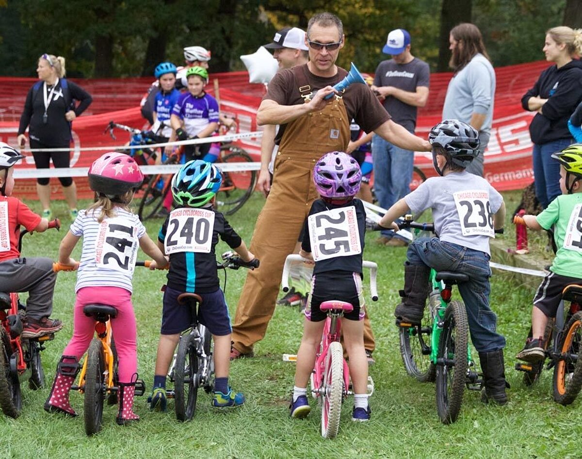 The start line of a junior race, official is holding a horn in his hand with the young racers looking up at him.